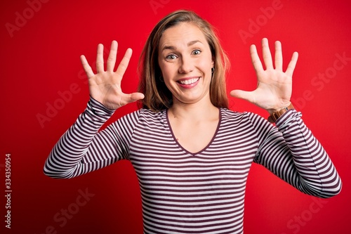 Young beautiful blonde woman wearing casual striped t-shirt over isolated red background showing and pointing up with fingers number ten while smiling confident and happy.