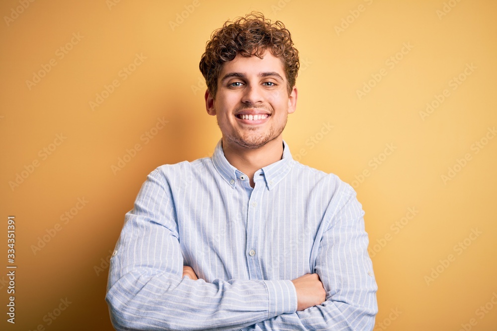 Young blond handsome man with curly hair wearing striped shirt over yellow background happy face smiling with crossed arms looking at the camera. Positive person. Young blond handsome man with curly hair wearing striped shirt over yellow background happy face smiling with crossed arms looking at the camera. Positive person.