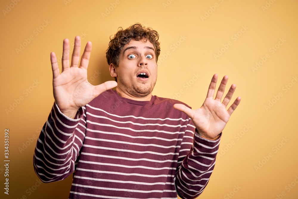 Young blond handsome man with curly hair wearing casual striped sweater afraid and terrified with fear expression stop gesture with hands, shouting in shock. Panic concept. Young blond handsome man with curly hair wearing casual striped sweater afraid and terrified with fear expression stop gesture with hands, shouting in shock. Panic concept.