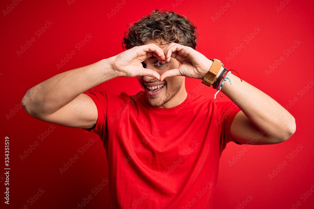 Young blond handsome man with curly hair wearing casual t-shirt over red background Doing heart shape with hand and fingers smiling looking through sign