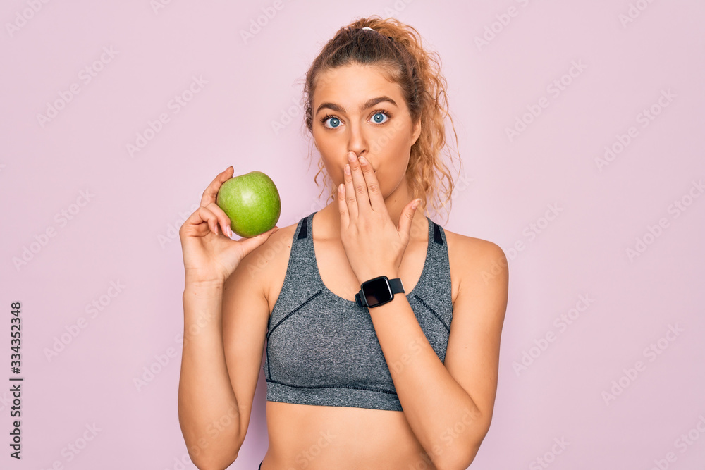 Young beautiful blonde sporty woman with blue eyes holding healthy green apple fruit cover mouth with hand shocked with shame for mistake, expression of fear, scared in silence, secret concept Young beautiful blonde sporty woman with blue eyes holding healthy green apple fruit cover mouth with hand shocked with shame for mistake, expression of fear, scared in silence, secret concept