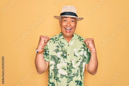 Middle age senior grey-haired man wearing summer hat and floral shirt on beach vacation celebrating surprised and amazed for success with arms raised and open eyes. Winner concept.