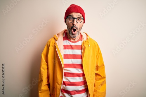 Middle age hoary man wearing glasses and rain coat standing over isolated white background afraid and shocked with surprise expression, fear and excited face.
