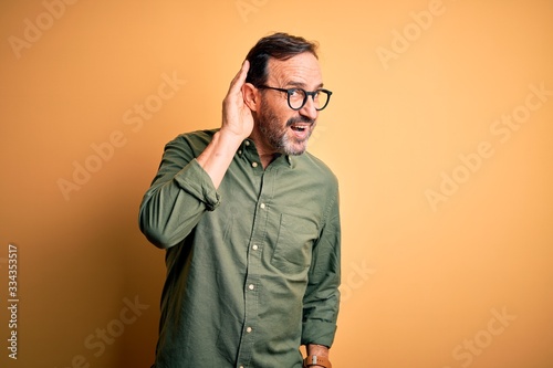 Middle age hoary man wearing casual green shirt and glasses over isolated yellow background smiling with hand over ear listening an hearing to rumor or gossip. Deafness concept.