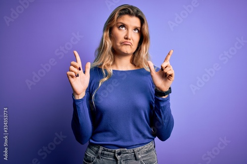Young beautiful blonde woman wearing casual t-shirt over isolated purple background Pointing up looking sad and upset, indicating direction with fingers, unhappy and depressed.