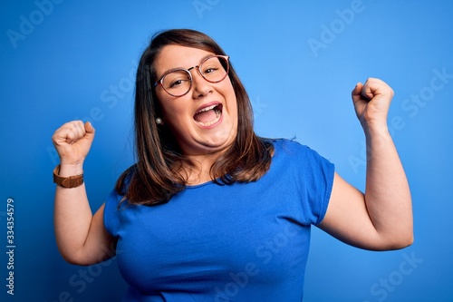 Beautiful brunette plus size woman wearing casual t-shirt over isolated blue background celebrating surprised and amazed for success with arms raised and open eyes. Winner concept.