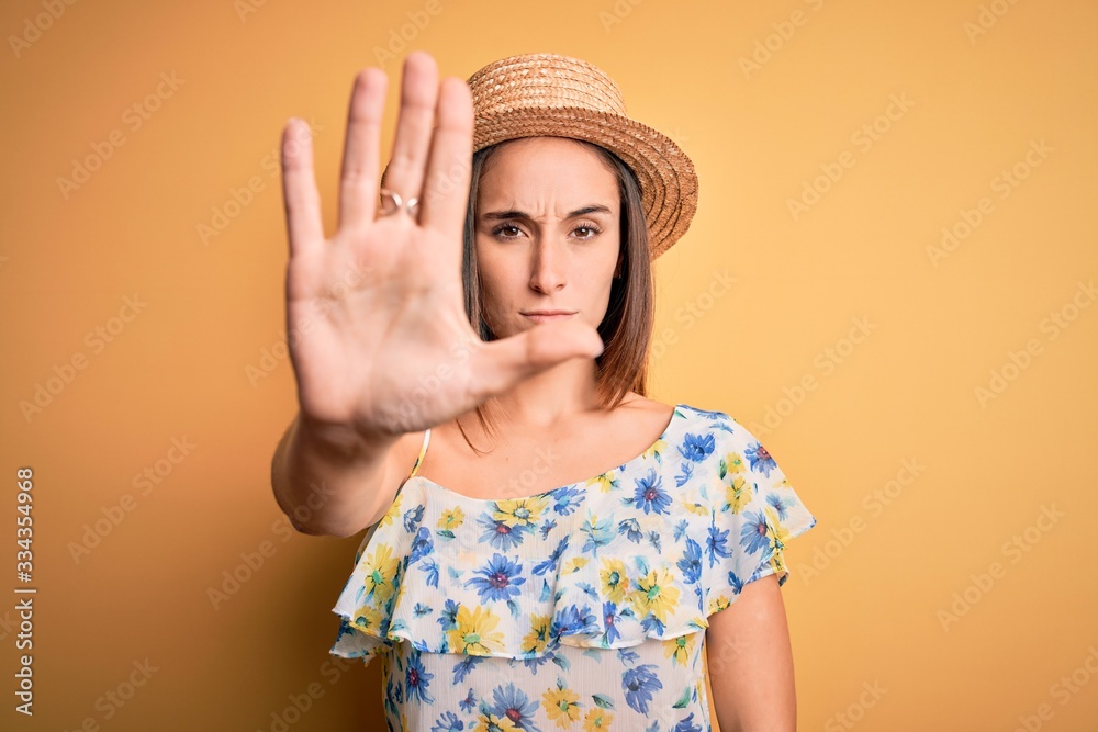 Young beautiful woman wearing casual t-shirt and summer hat over isolated yellow background doing stop sing with palm of the hand. Warning expression with negative and serious gesture on the face. Young beautiful woman wearing casual t-shirt and summer hat over isolated yellow background doing stop sing with palm of the hand. Warning expression with negative and serious gesture on the face.