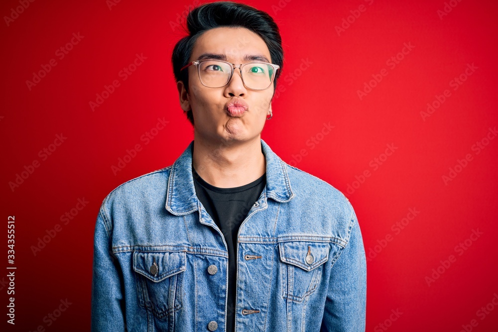 Young handsome chinese man wearing denim jacket and glasses over red background making fish face with lips, crazy and comical gesture. Funny expression. Young handsome chinese man wearing denim jacket and glasses over red background making fish face with lips, crazy and comical gesture. Funny expression.
