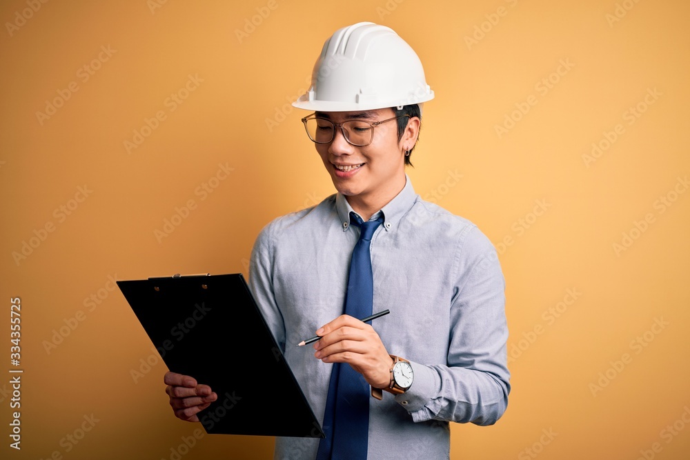 Young handsome chinese engineer man wearing safety helmet holding clipboard with a happy face standing and smiling with a confident smile showing teeth Young handsome chinese engineer man wearing safety helmet holding clipboard with a happy face standing and smiling with a confident smile showing teeth