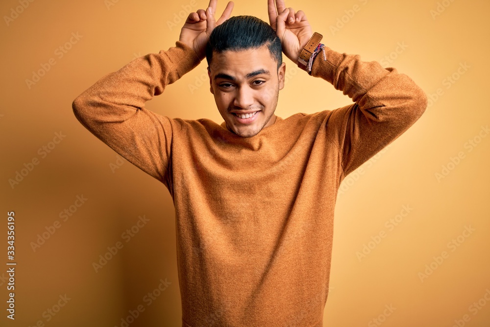 Young brazilian man wearing casual sweater standing over isolated yellow background Posing funny and crazy with fingers on head as bunny ears, smiling cheerful Young brazilian man wearing casual sweater standing over isolated yellow background Posing funny and crazy with fingers on head as bunny ears, smiling cheerful