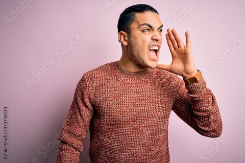 Young brazilian man wearing casual sweater standing over isolated pink background shouting and screaming loud to side with hand on mouth. Communication concept.