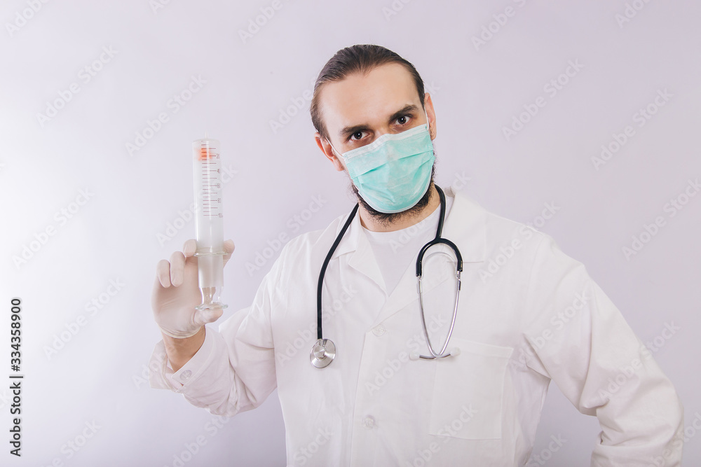 A doctor in a white coat, a medical mask and gloves holds a large syringe. Studio photo on a white background