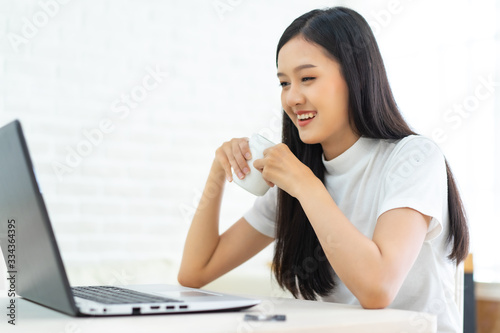 Young Asian  woman relaxing and drinking cup of hot coffee or tea using laptop computer on  winter day in the bedroom.woman checking social apps and working.Work at home concept. © anon