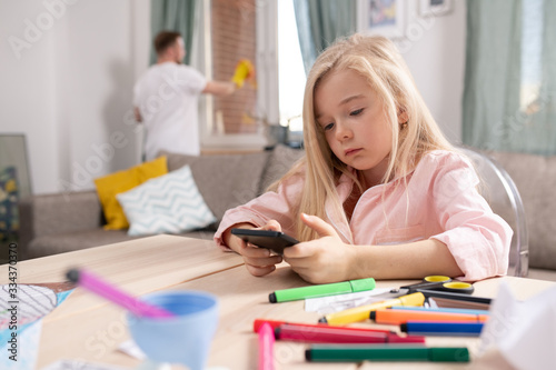 Little restful blond girl scrolling in smartphone while sitting in living-room