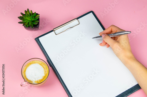 Tablet with white flattery next to which stands a green plant, hand holds a pen and writes on a sheet in the other hand a cup with a coffe photo