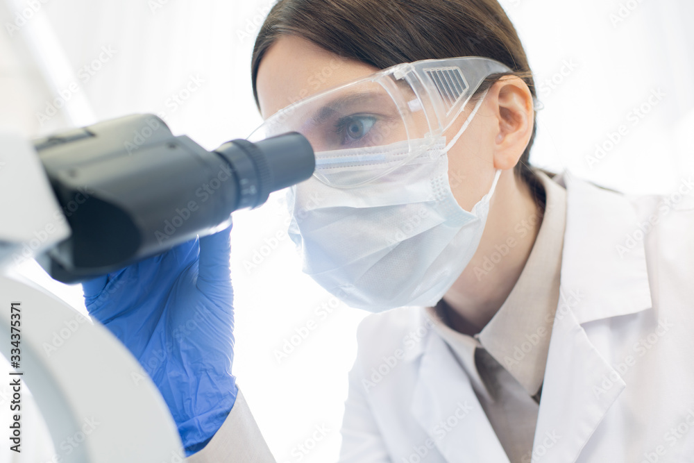 Female researcher in gloves, mask and protective eyewear bending over microscope Female researcher in gloves, mask and protective eyewear bending over microscope