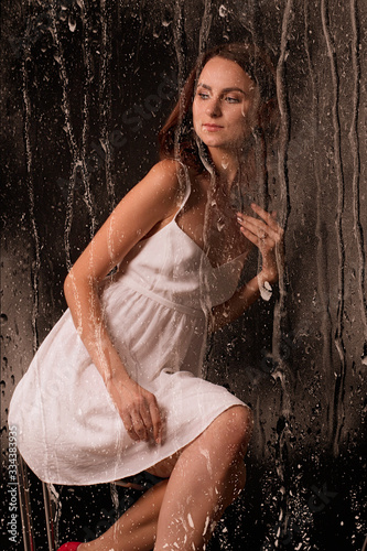 water drops flow like rain on glass, special effects on the set, in the background a beautiful and sexy young brunette girl in a white short dress sits in a high chair on a black background