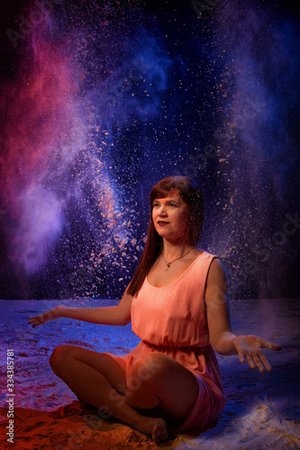 Beautiful woman with long brunette hair in pink dress with colored lights and flour around. Portrait of girl and female model posing in photoshoot with flour at studio with black background