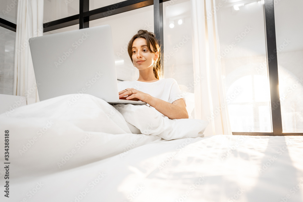 Woman working on a laptop, lying in the beautiful white and sunny bedroom. Work from home at cozy atmosphere concept