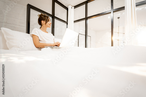 Woman working on a laptop, lying in the beautiful white and sunny bedroom. Work from home at cozy atmosphere concept