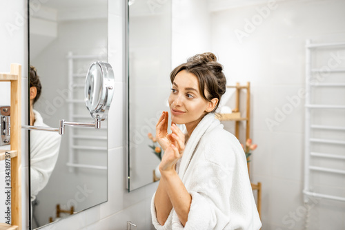 Young woman in bathrobe taking care of her skin, looking at the mirror in the bathroom. Facial skin care and wellness concept