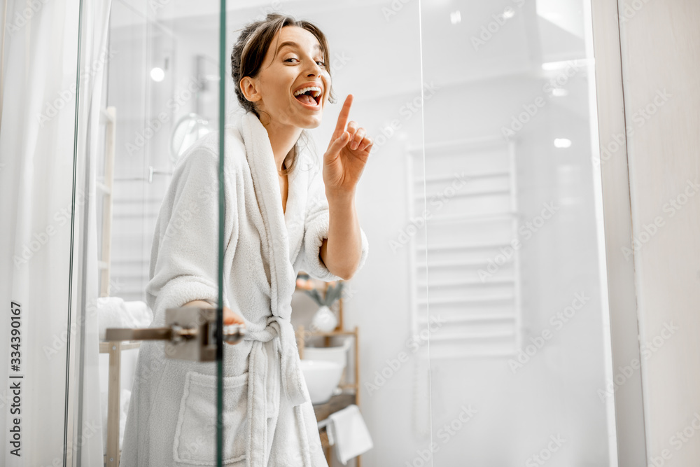 Young and joyful woman in bathrobe going to the bath, closing a glass doors at home - obrazy, fototapety, plakaty 