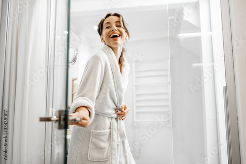 Young and joyful woman in bathrobe going to the bath, closing a glass doors at home photo