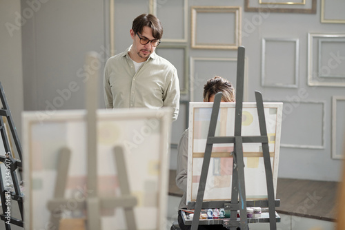 Serious teacher of professional painting course standing by one of his students