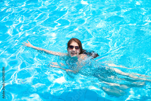 Beautiful joyful girl with long hair in sunglasses swims in the pool on a summer day. Travel, resort