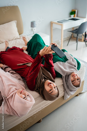 three veiled women lay down and hands raised on the bed while taking a selfie together with camera smartphone