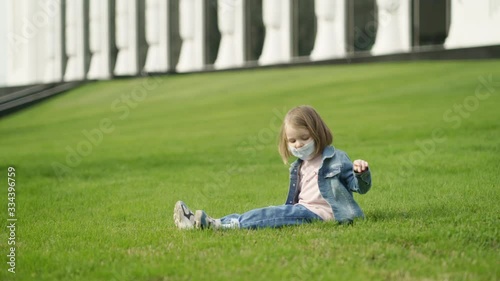 White little child girl wear protection mask and sits on the grass. Spring windy Quarantine day 