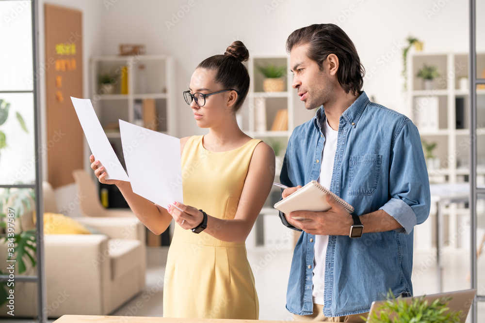 Young female office worker comparing two papers while consulting with colleague Young female office worker comparing two papers while consulting with colleague