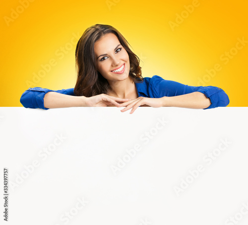 Portrait of smiling attractive woman holding blank sign board with copy space, isolated over yellow orange background. Success in business concept. Brunette model in blue clothing at studio picture.