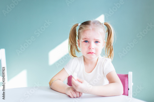 Little blonde girl with two ponytales is crying sitting by the white table with tears on her face. Copyspace. photo