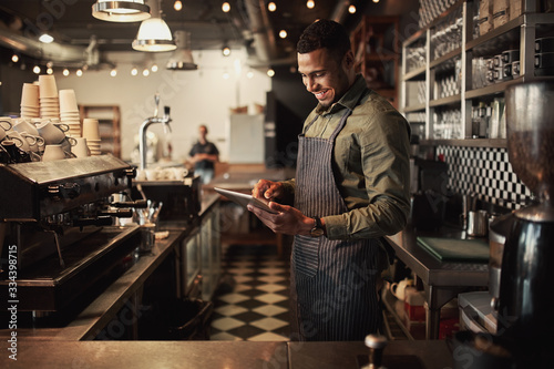 Cheerful young afro-american cafe owner wearing black striped apron using digital tablet