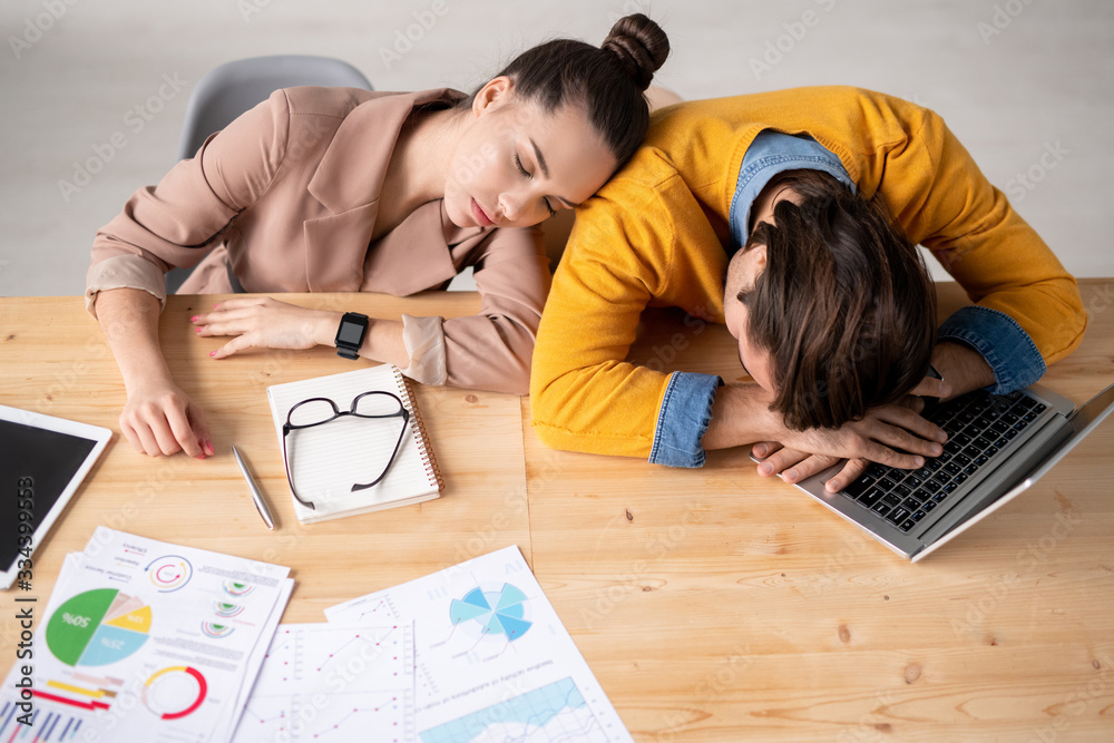 Two young exhausted brokers napping by desk after hard working day in office Two young exhausted brokers napping by desk after hard working day in office