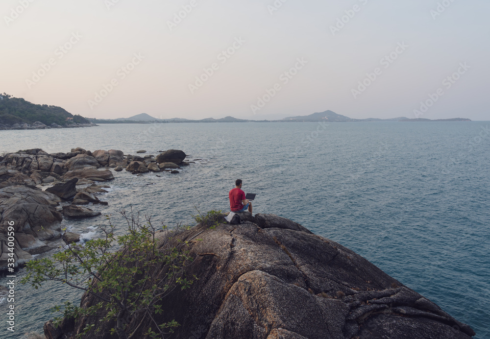 A man sits on a rock by the sea and works on laptop