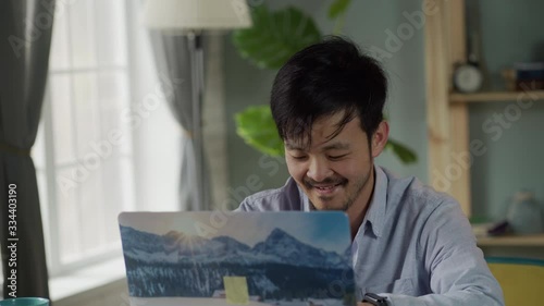 Work from Home. Cheerful young Korean man with a mustache and beard sits at a Desk, intently working on laptop and smiling in a comfortable home environment.