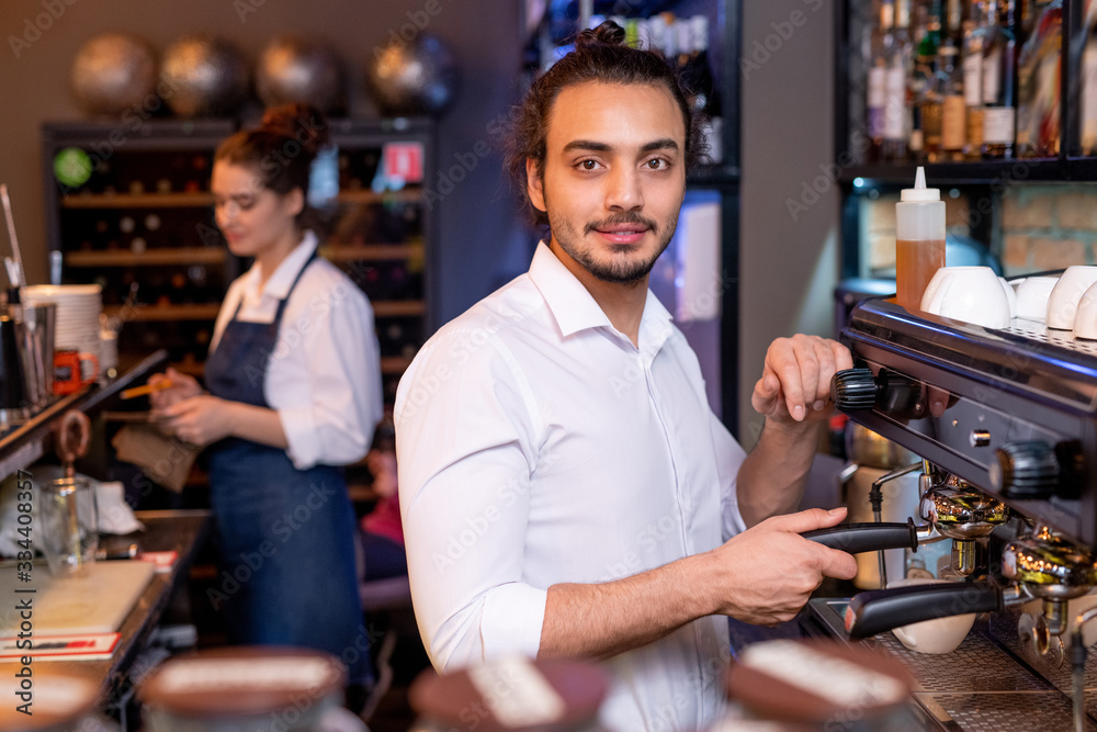Young handsome waiter in white shirt preparing cappuccino by coffee machine