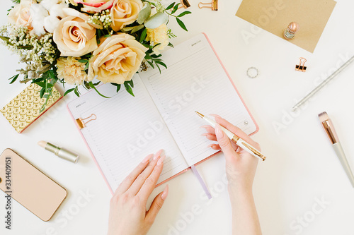 Female hands wtite in pink opened notebook on white background. Tender mood and blogging concept with beautiful roses bouquet and accesoires in flat lay style photo