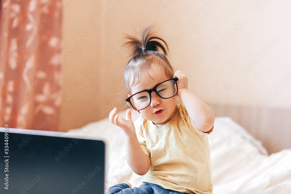 Little happy funny black-haired girl using laptop in glasses on bed. boy sitting ubfocused glasses in focus. Concept problem of ophthalmology correction of myopia Little happy funny black-haired girl using laptop in glasses on bed. boy sitting ubfocused glasses in focus. Concept problem of ophthalmology correction of myopia
