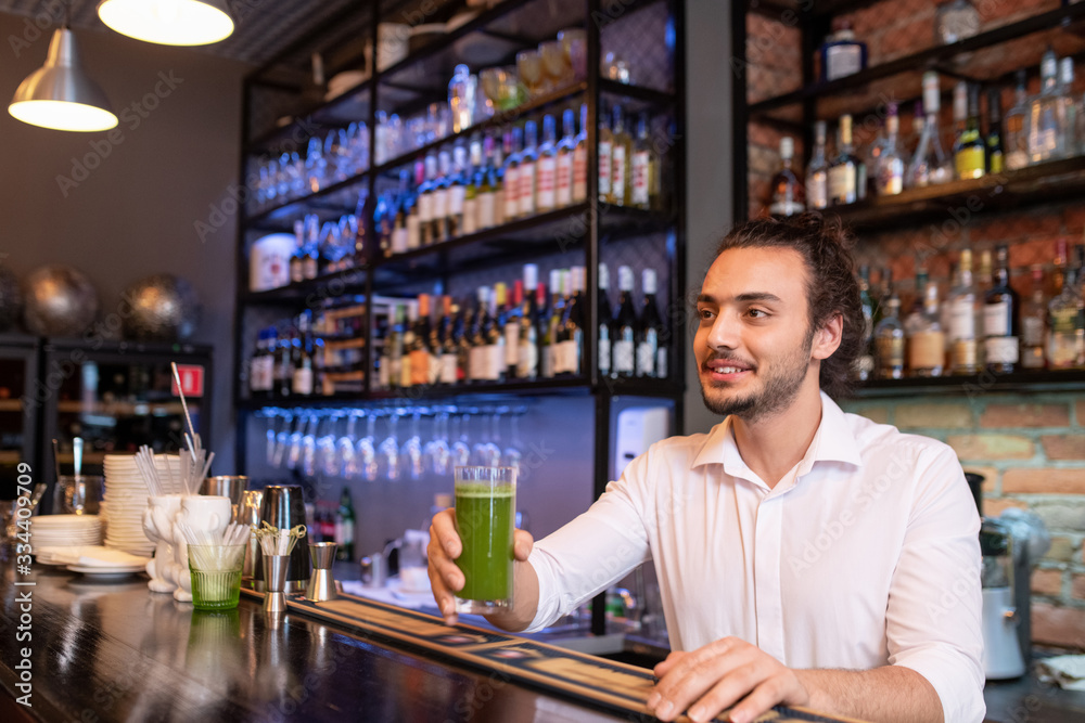 Young smiling barman holding glass of green salary cocktail or smoothie Young smiling barman holding glass of green salary cocktail or smoothie