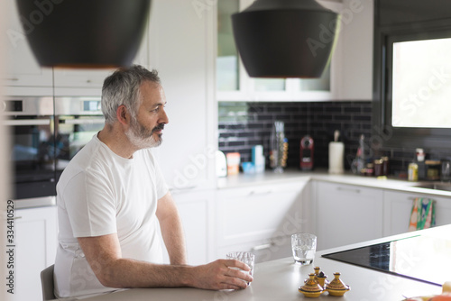 Portrait of bearded man sitting at the kitchen holding a glass of water while looking aside pensive