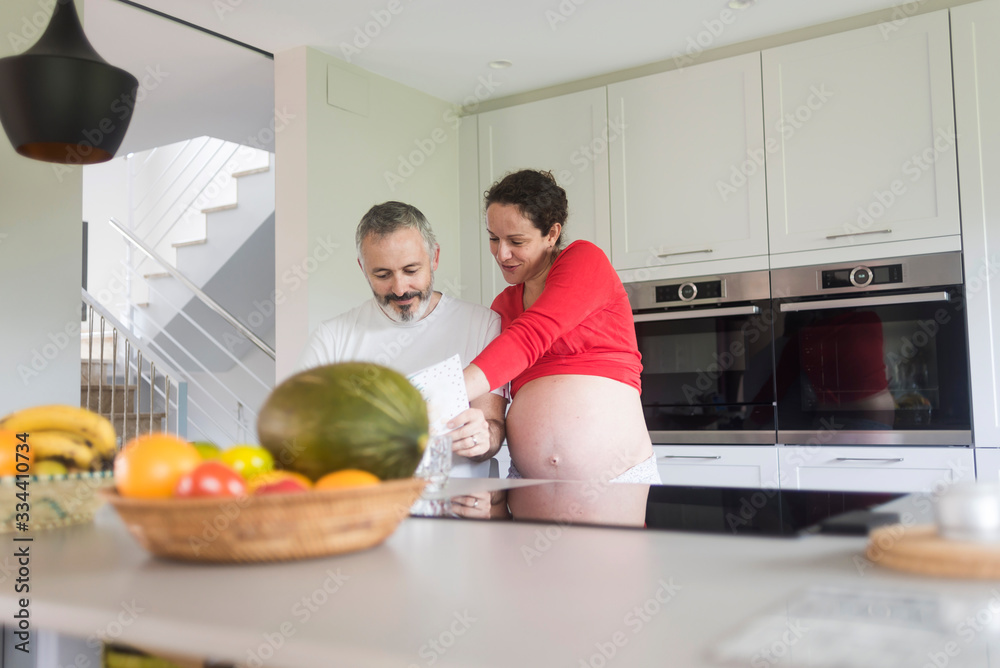 Happy pregnant woman with her husband sitting at home. Smiling couple consulting a book at the kitchen. Happy pregnant woman with her husband sitting at home. Smiling couple consulting a book at the kitchen.