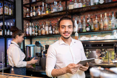 Young cheerful waiter with touchpad standing in front of camera