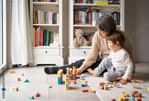 Little toddler boy playing wooden building blocks with grandmother at home. Family spending time together at quarantine period during pandemic. Bright lifestyle.