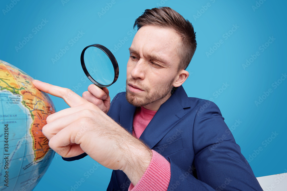 Serious guy squinting one eye while looking at globe through magnifying glass Serious guy squinting one eye while looking at globe through magnifying glass