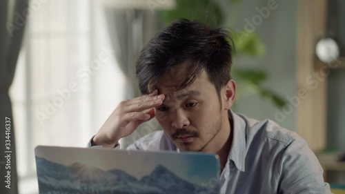 Work from home. Sick and tired young Korean man with a mustache and a beard sits at a desk, working on a laptop at home and coughs