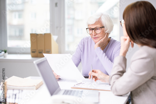Serious aged woman looking through insurance form while sitting by desk