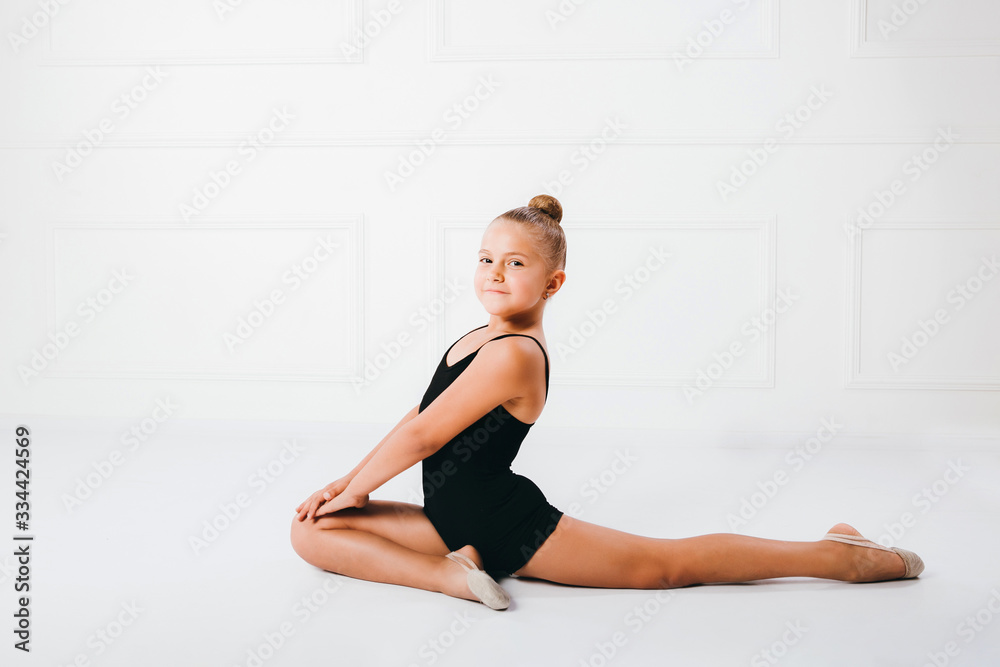 Flexible little girl gymnast sitting in acrobatic pose on white background.